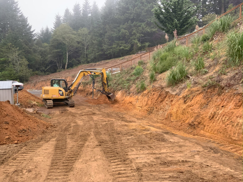 Excavator digging into a hillside, creating a cleared dirt path surrounded by trees and vegetation.