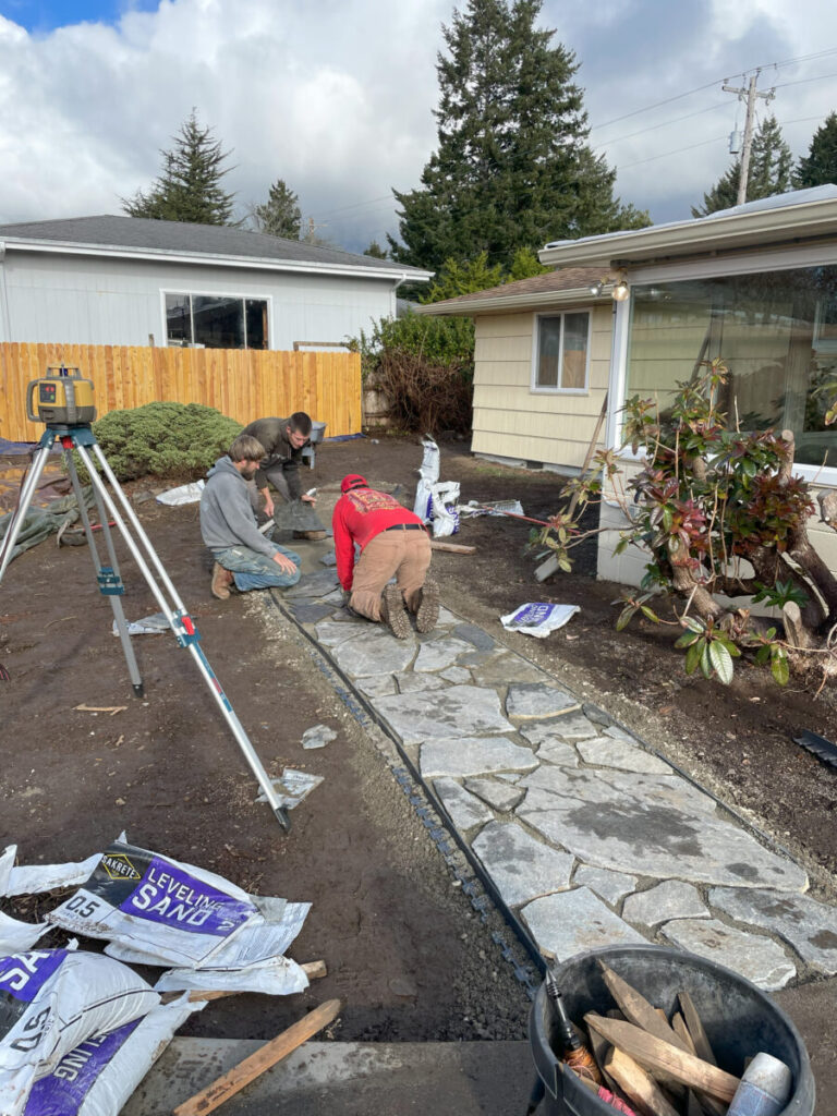 Three people are installing stone pavers to build a walkway in a residential backyard on a partly cloudy day.
